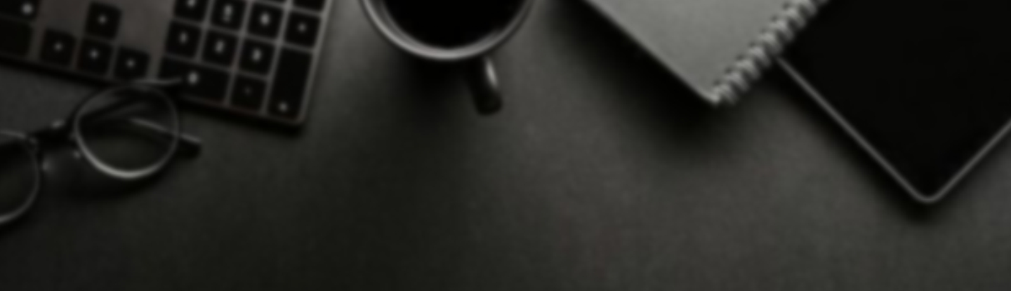 Imagem de cima de uma mesa de trabalho, escura, exibindo partes de um óculos, teclado, xícara, bloco de anotações e tablet.