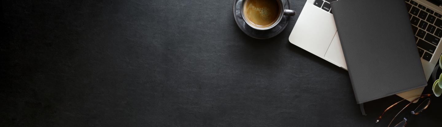 Imagem de cima de uma mesa de trabalho, escura, exibindo partes de um notebook, óculos, caderno de anotações e café.