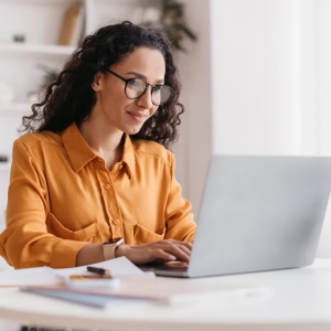 Foto de uma mulher branca de óculos de grau e cabelos escuros cacheados, vestindo uma camisa amarela escura usando um notebook claro sobre uma mesa clara.