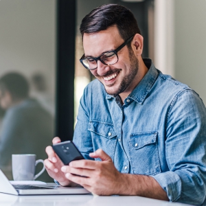 Foto de um homem branco com barba, camisa jeans e óculos de grau usando um smartphone e sorrindo.