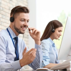 Foto de um homem e uma mulher brancos vestindo camisa social azul clara usando computadores e um fone de ouvido com microfone.