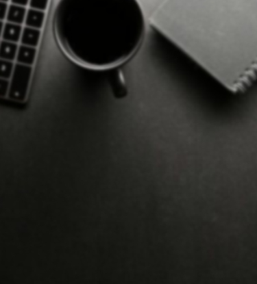 Imagem de cima de uma mesa de trabalho, escura, exibindo partes de um óculos, teclado, xícara, bloco de anotações e tablet.