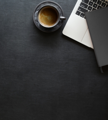 Imagem de cima de uma mesa de trabalho, escura, exibindo partes de um notebook, óculos, caderno de anotações e café.