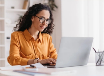 Foto de uma mulher branca de óculos de grau e cabelos escuros cacheados, vestindo uma camisa amarela escura usando um notebook claro sobre uma mesa clara.