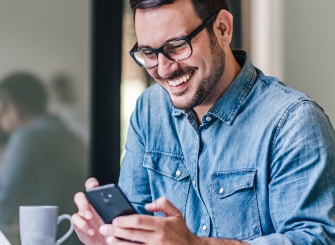 Foto de um homem branco com barba, camisa jeans e óculos de grau usando um smartphone e sorrindo.