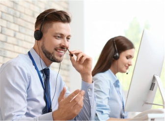 Foto de um homem e uma mulher brancos vestindo camisa social azul clara usando computadores e um fone de ouvido com microfone.