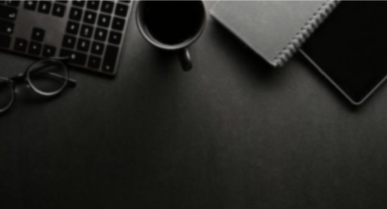 Imagem de cima de uma mesa de trabalho, escura, exibindo partes de um óculos, teclado, xícara, bloco de anotações e tablet.
