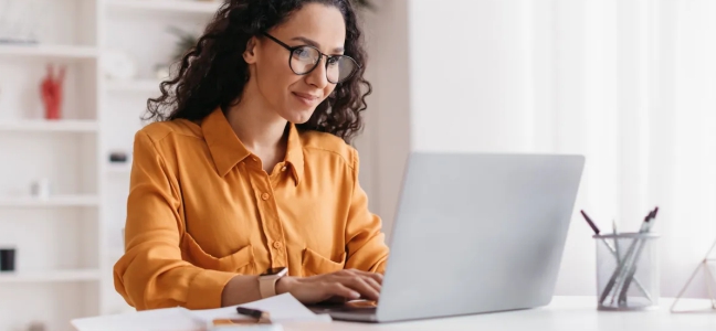 Foto de uma mulher branca de óculos de grau e cabelos escuros cacheados, vestindo uma camisa amarela escura usando um notebook claro sobre uma mesa clara.