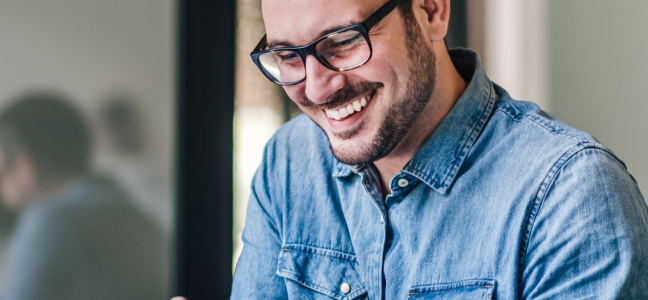 Foto de um homem branco com barba, camisa jeans e óculos de grau usando um smartphone e sorrindo.