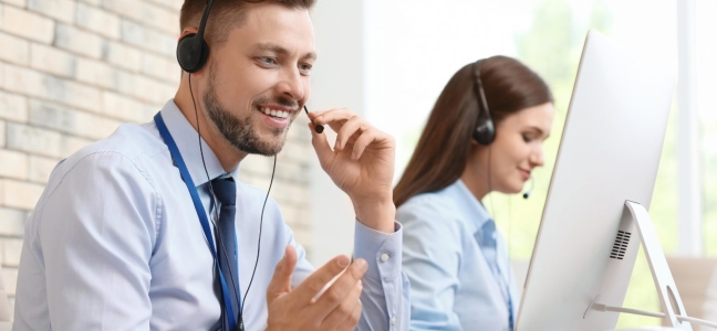 Foto de um homem e uma mulher brancos vestindo camisa social azul clara usando computadores e um fone de ouvido com microfone.